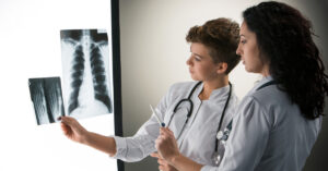 Two female doctors in white coats examining an X-ray of a human chest on a light board.